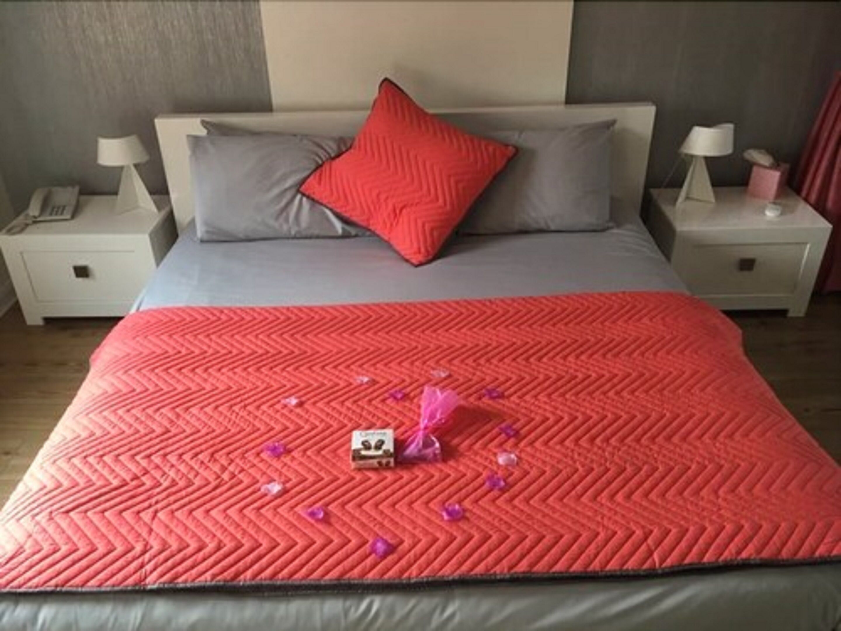 Uma cama com um edredom vermelho, um travesseiro vermelho e um travesseiro cinza. Existem duas mesas de cabeceira, uma com luminárias e um telefone.