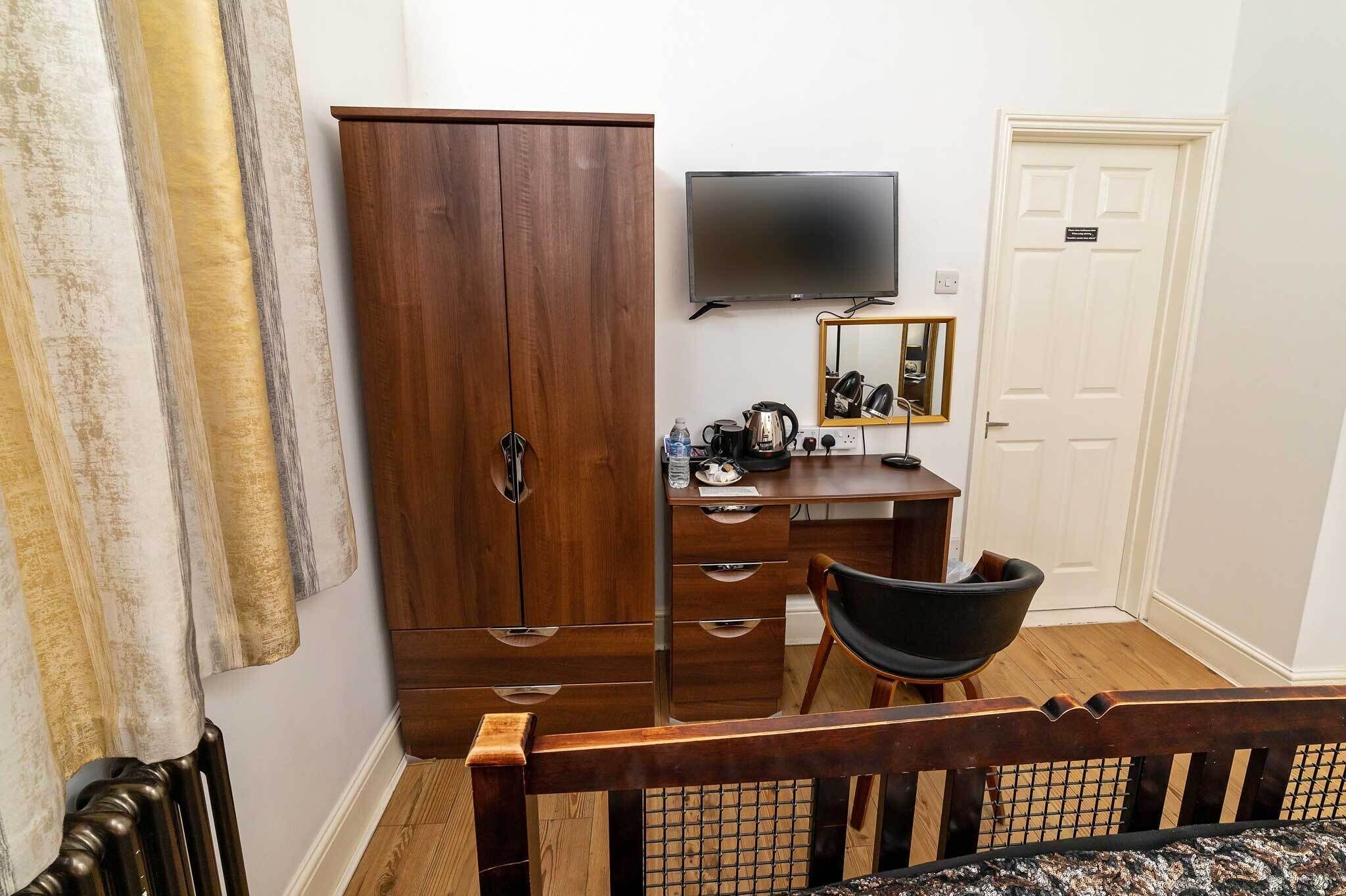 A bedroom with a wooden wardrobe, a desk with a mirror, a television, and a bed with a headboard.