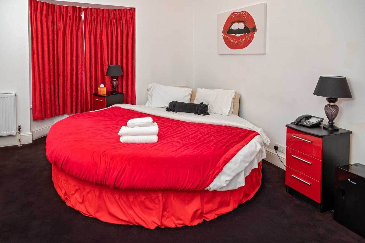 A bedroom with a large bed, red curtains, a bedside table with a lamp, and a wall art piece.