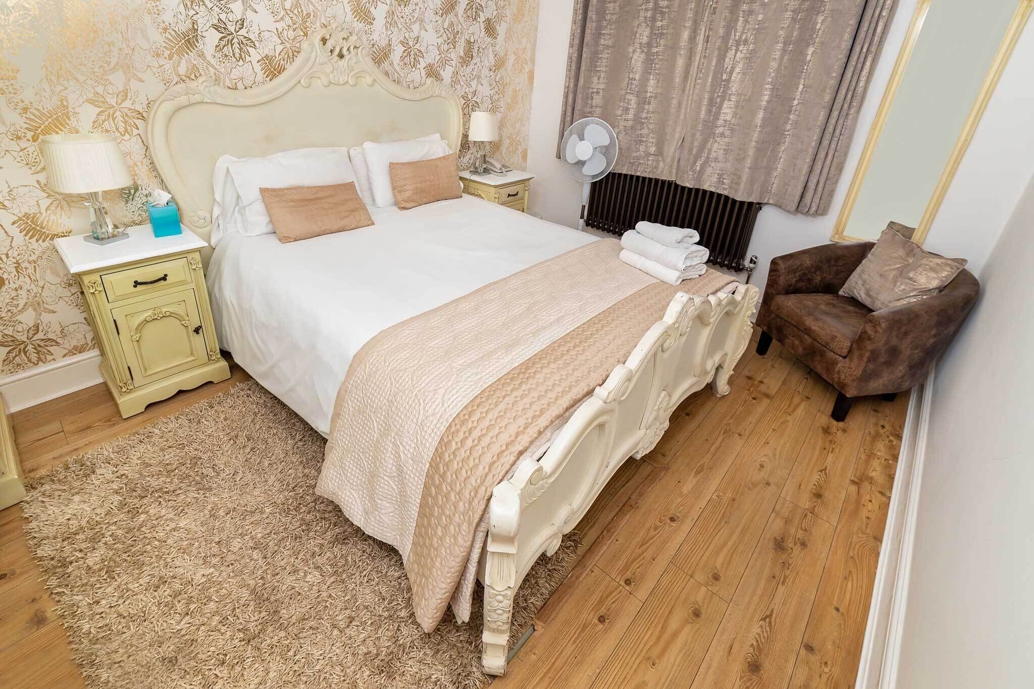 A neatly made bed with a headboard, a bedside table with a lamp, a chair, and a wall-mounted fan.