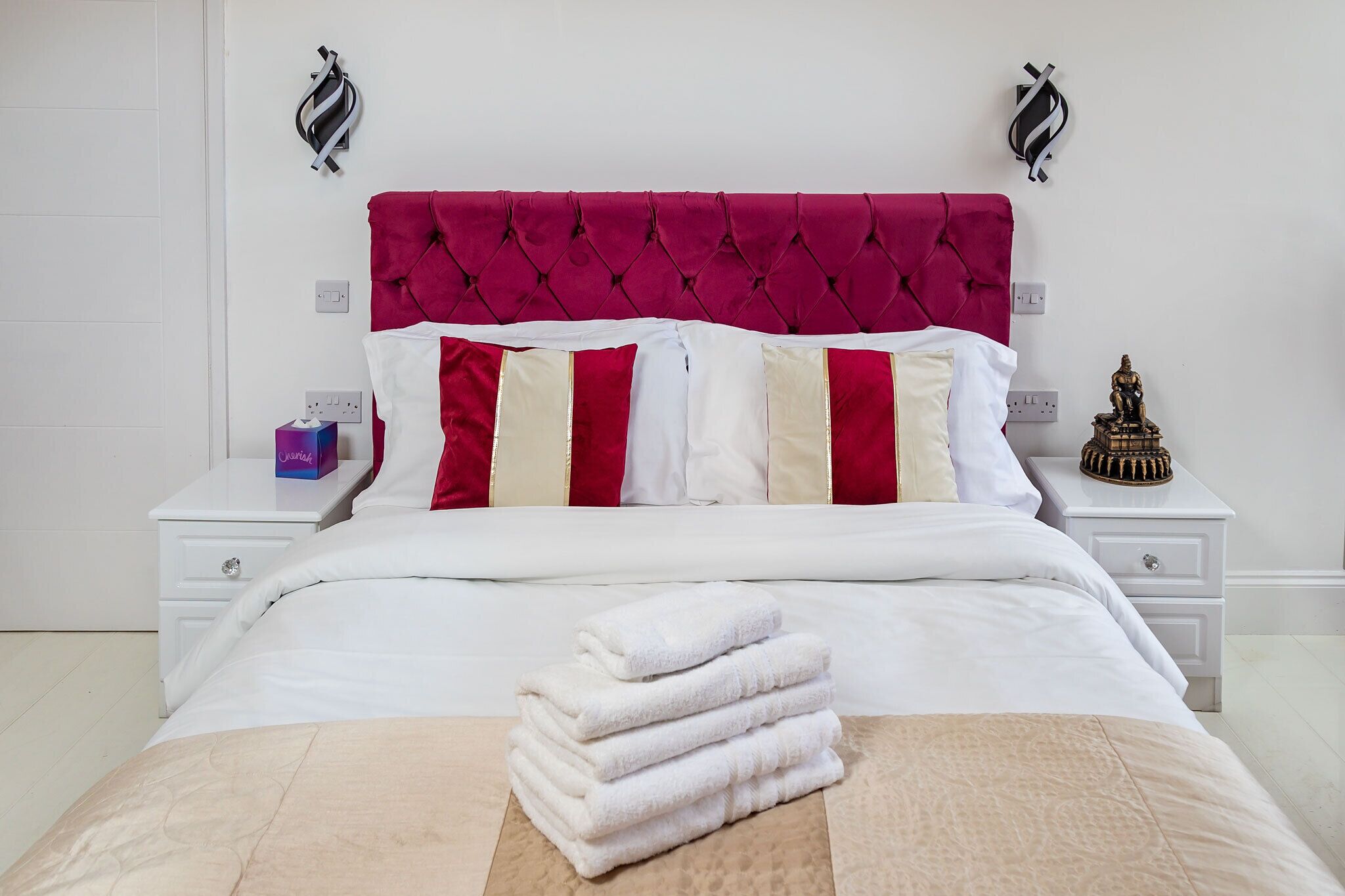 Una cama bien tendida con cabecera roja, ropa de cama blanca y una pila de toallas blancas al pie.