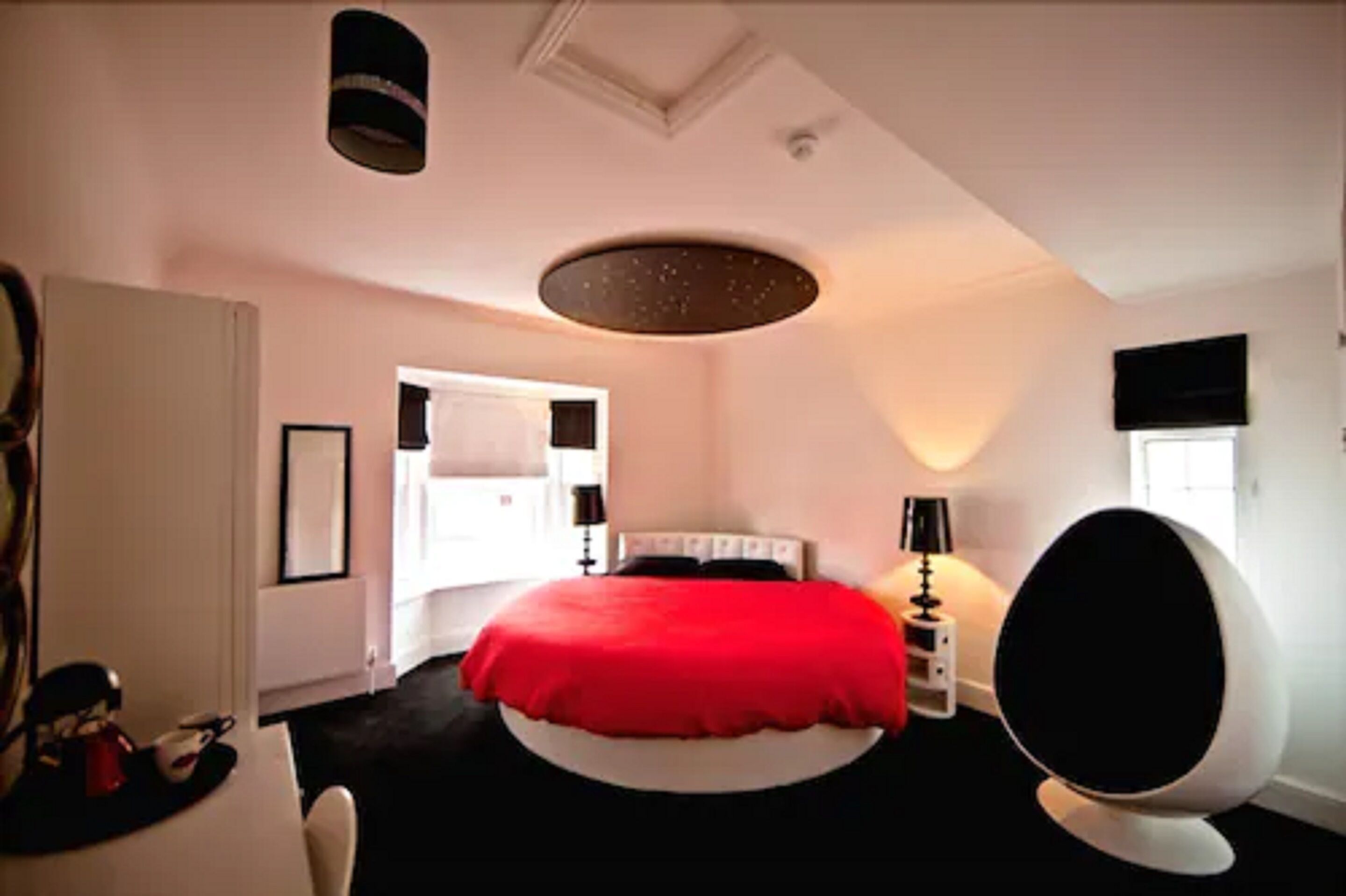 A bedroom with a red bedspread, a round chair, a desk with a chair, and a television on the wall.