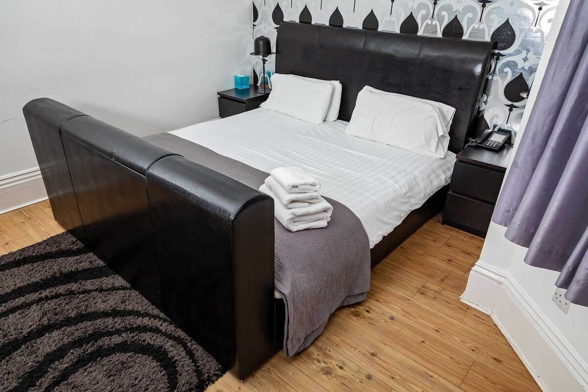 A neatly made bed with a folded white towel, a black headboard, and a bedside table with a lamp and a phone.