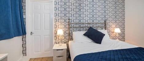 Un dormitorio con un cabecero estampado, una cama con colcha azul, dos lámparas de noche y una mesita de noche blanca.