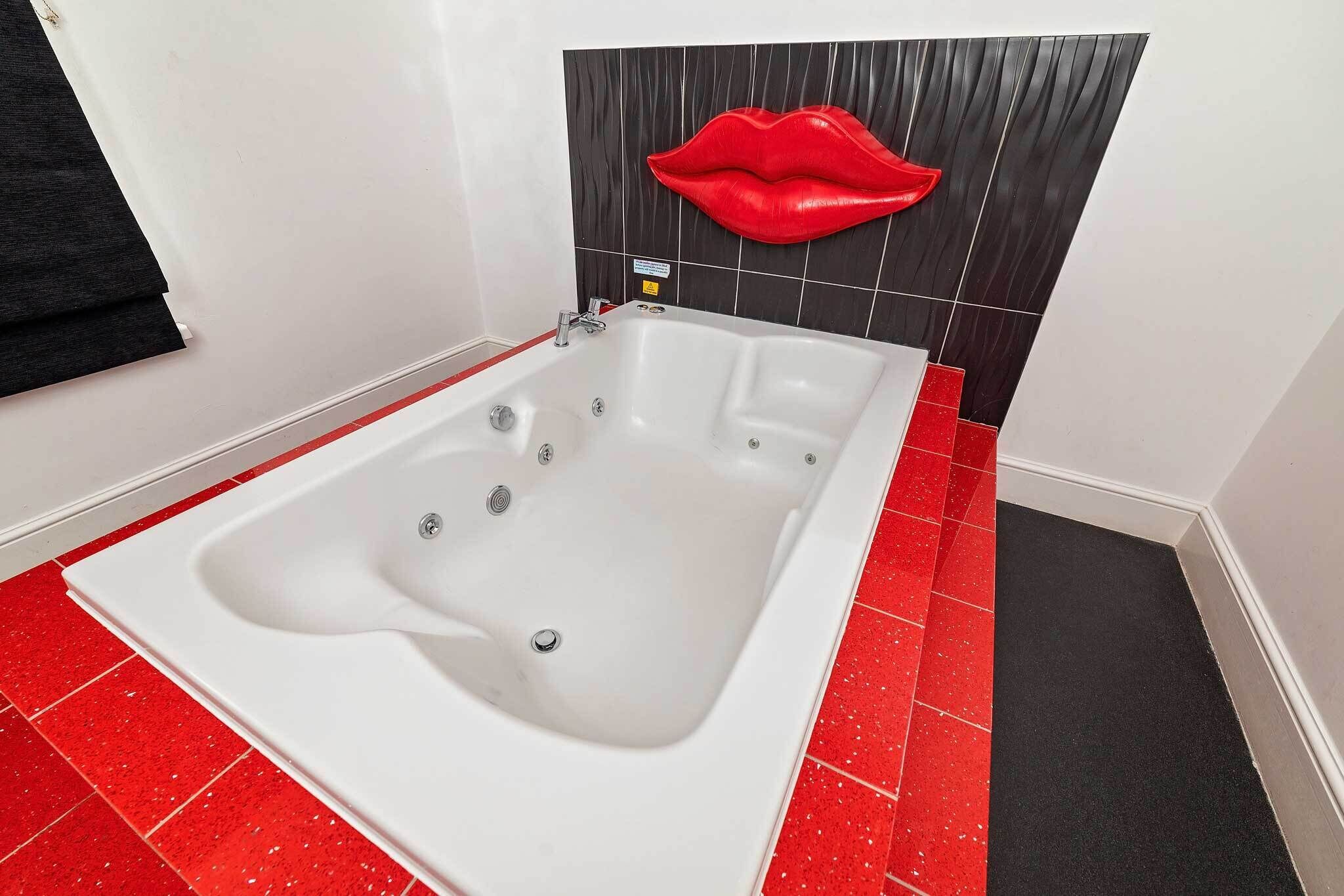 A modern bathroom with a large bathtub, red and black tiled floor, and a wall-mounted red lips decoration.