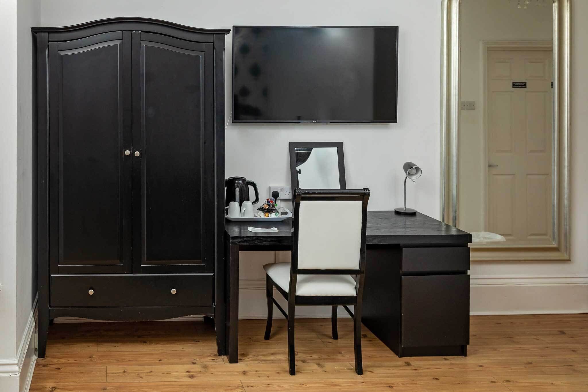 Een kamer met een donker houten bureau, een witte stoel, een flatscreen televisie, een spiegel en een kledingkast.
