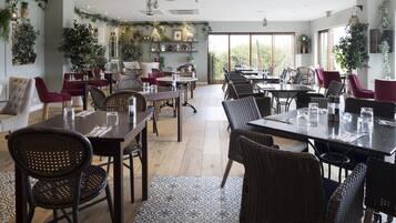 Un comedor amplio con varias mesas y sillas, rodeado de ventanas grandes y plantas de interior.