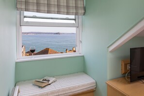 A small room with a window overlooking the sea, a bed with a book and glasses, and a TV on a wooden stand.
