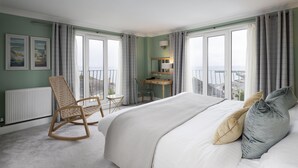 A bedroom with a large bed, a desk, a rocking chair, and a view of the sea through large windows. - Hotel Penzance Harbour View (Penzance)