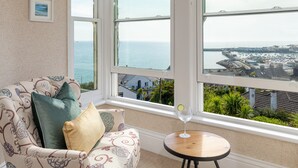 A room with a large bay window overlooking a coastal view, a patterned sofa, and a small round table with a glass of water. - Hotel Penzance Harbour View (Penzance)