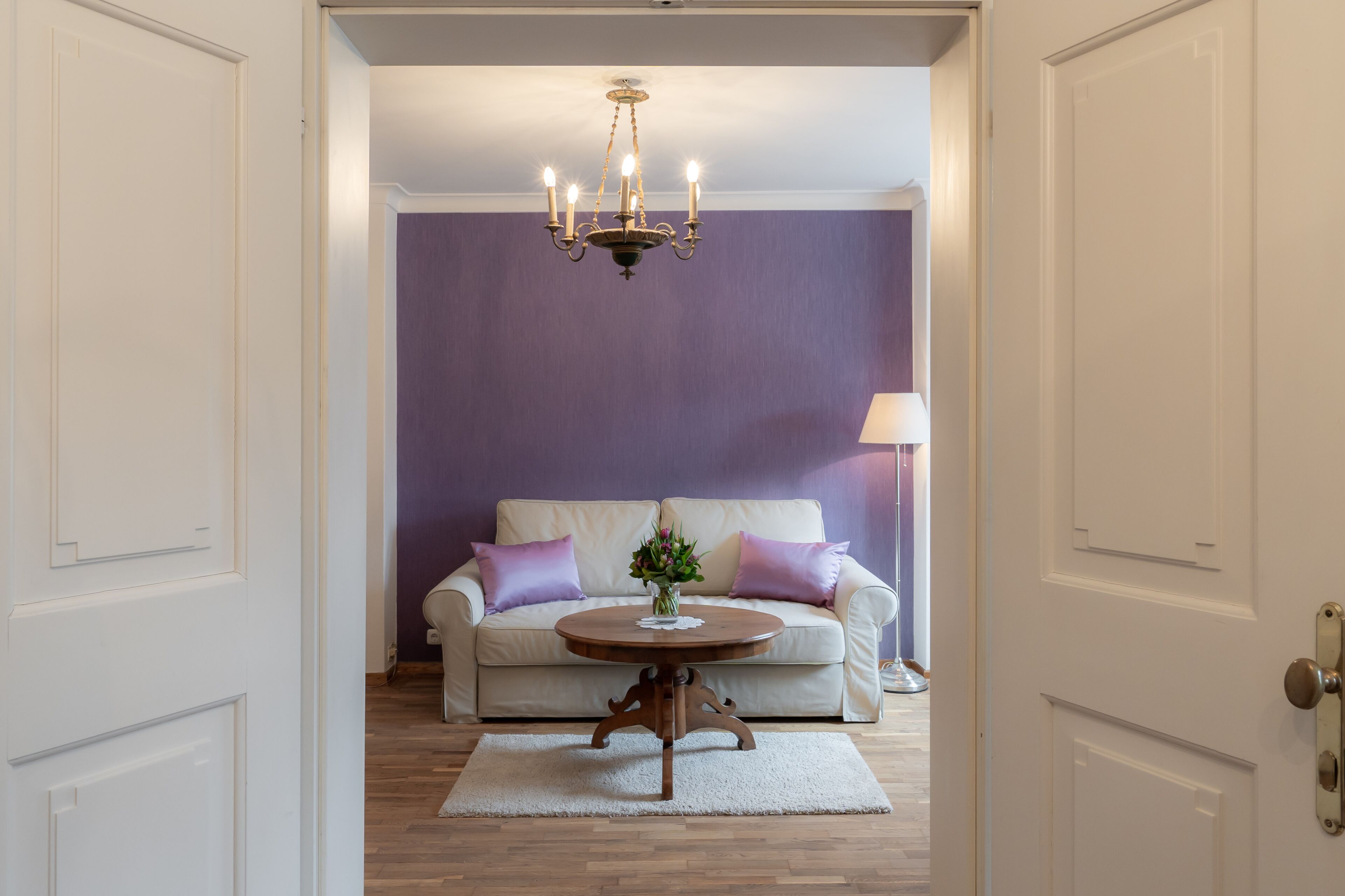 Una habitación con una puerta blanca, un sofá blanco con cojines morados, una mesa de centro redonda de madera y un candelabro.
