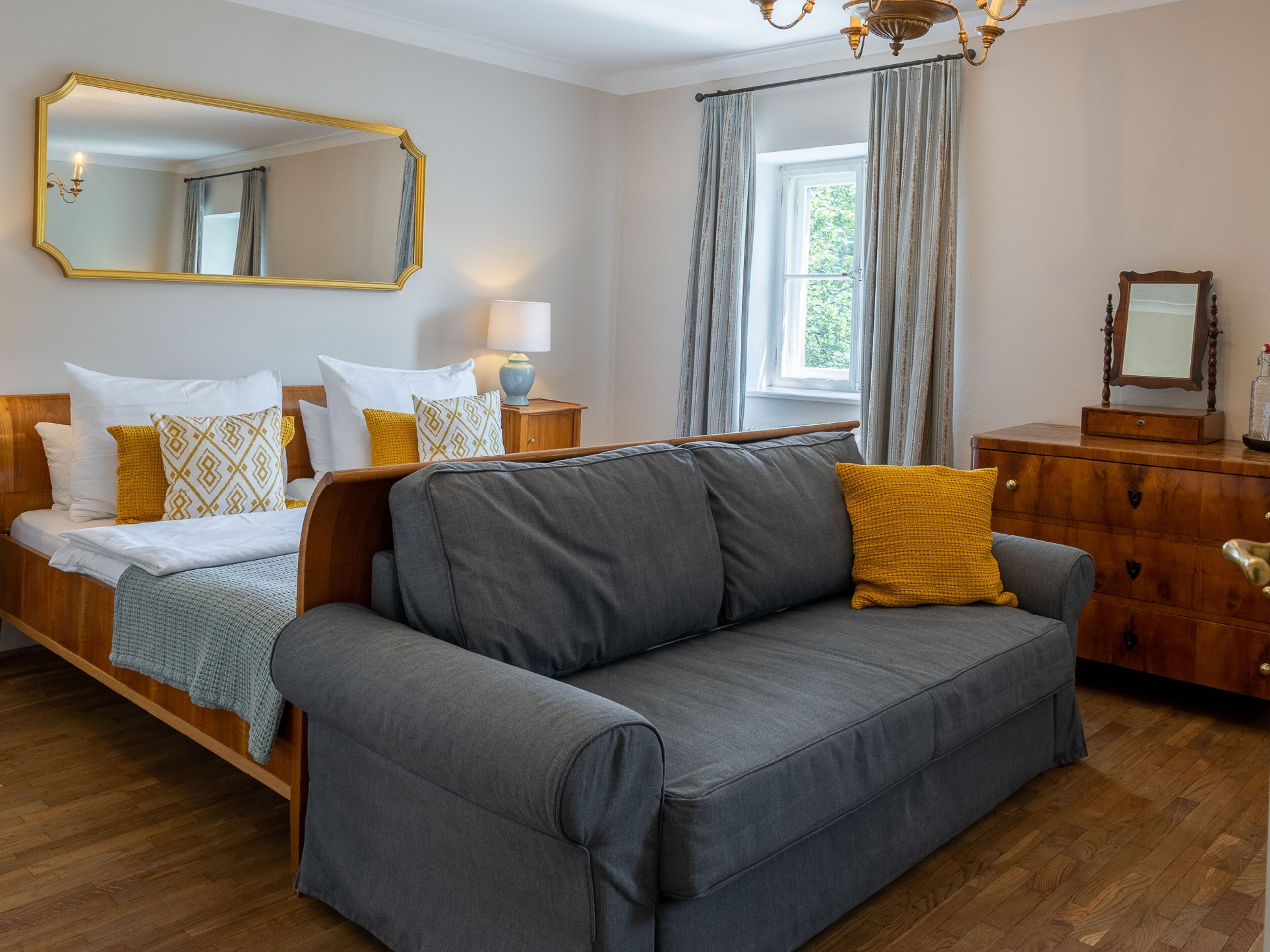 A modern hotel room with a grey sofa, wooden headboard, and a large mirror.