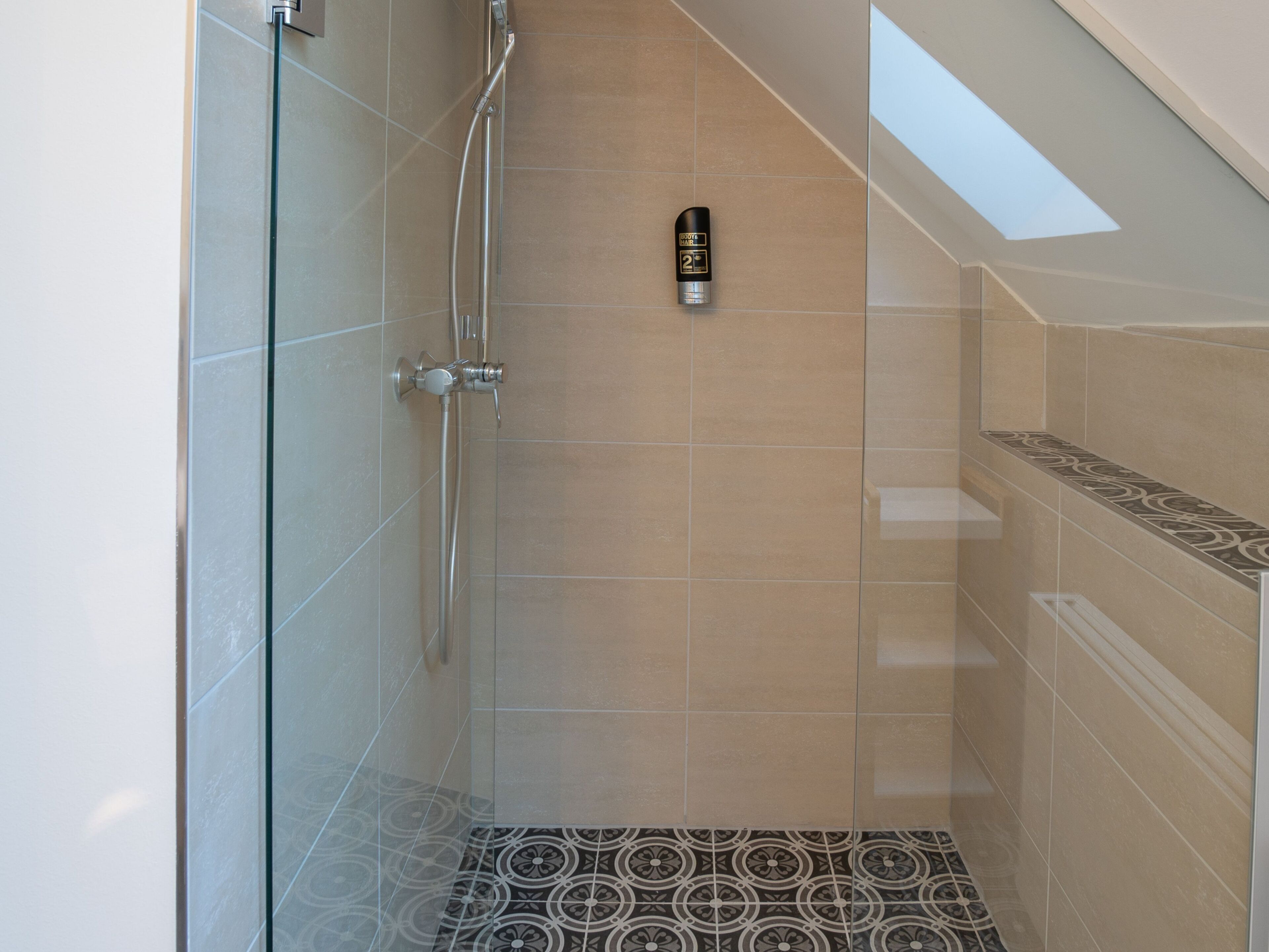 Eine Dusche mit Glastür, Duschbrausehandstück und einer Shampoo-Flasche an der Wand.