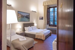 Een hotelkamer met twee bedden, een nachtkastje met een lamp, een schilderij van een brug en een raam met gordijnen.