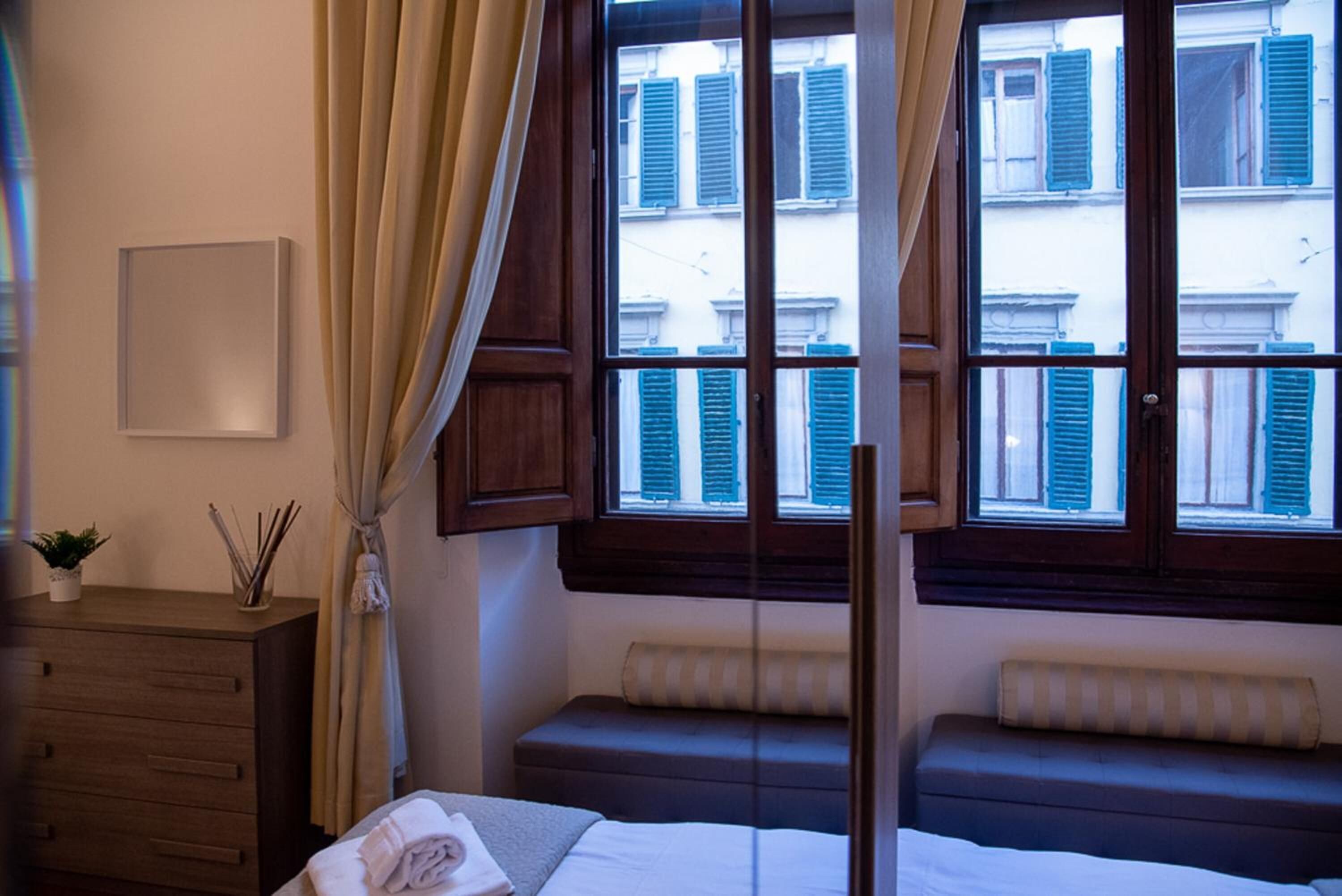 A hotel room with a bed, a bench, a dresser, a potted plant, and a window with blue shutters.