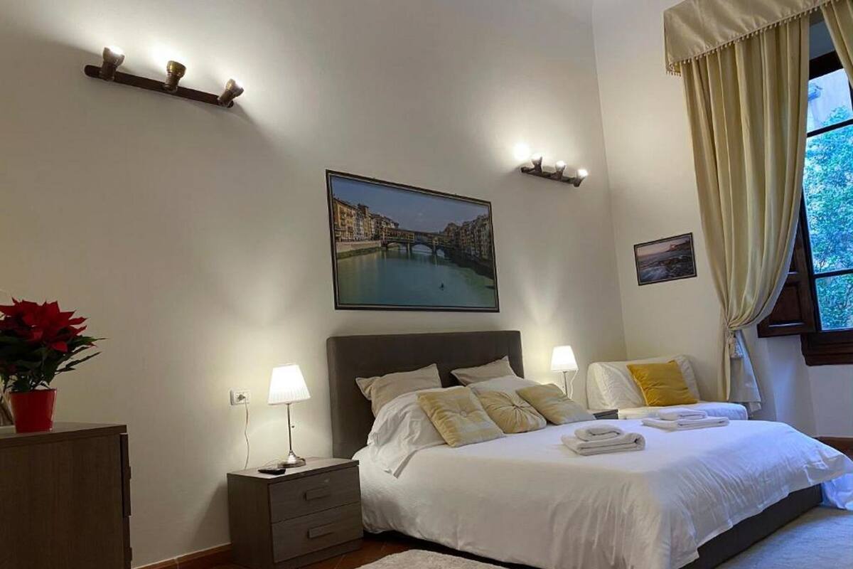 Une chambre à coucher bien rangée, avec un grand lit, des lampes de chevet, une table de nuit, une plante en pot rouge et un tableau représentant un paysage urbain accroché au mur.