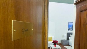 Ein Hotelzimmer mit einer Holztür mit dem Schild "Giotto", einem weißen Bett, einem kleinen weißen Tisch mit einer roten Blumenvase, einem grauen Nachttisch und einer weißen Lampe.