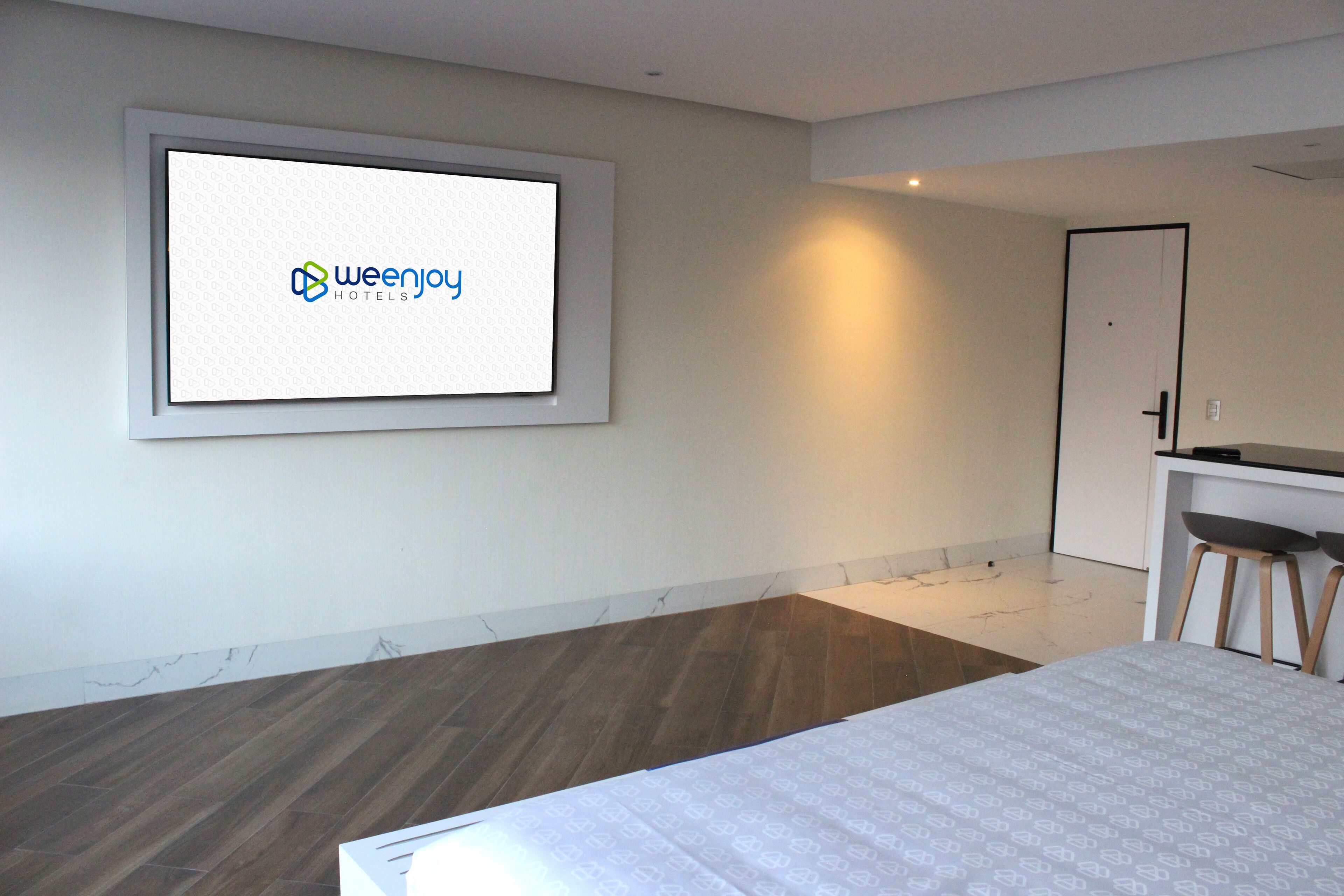 A modern hotel room with a large bed, a desk with two chairs, a small kitchenette, and a framed screen displaying the Weenjoy Hotels logo.