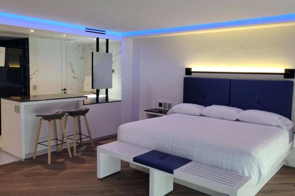 A modern bedroom with a large bed, a small desk with stools, and a bathroom visible through an open door.