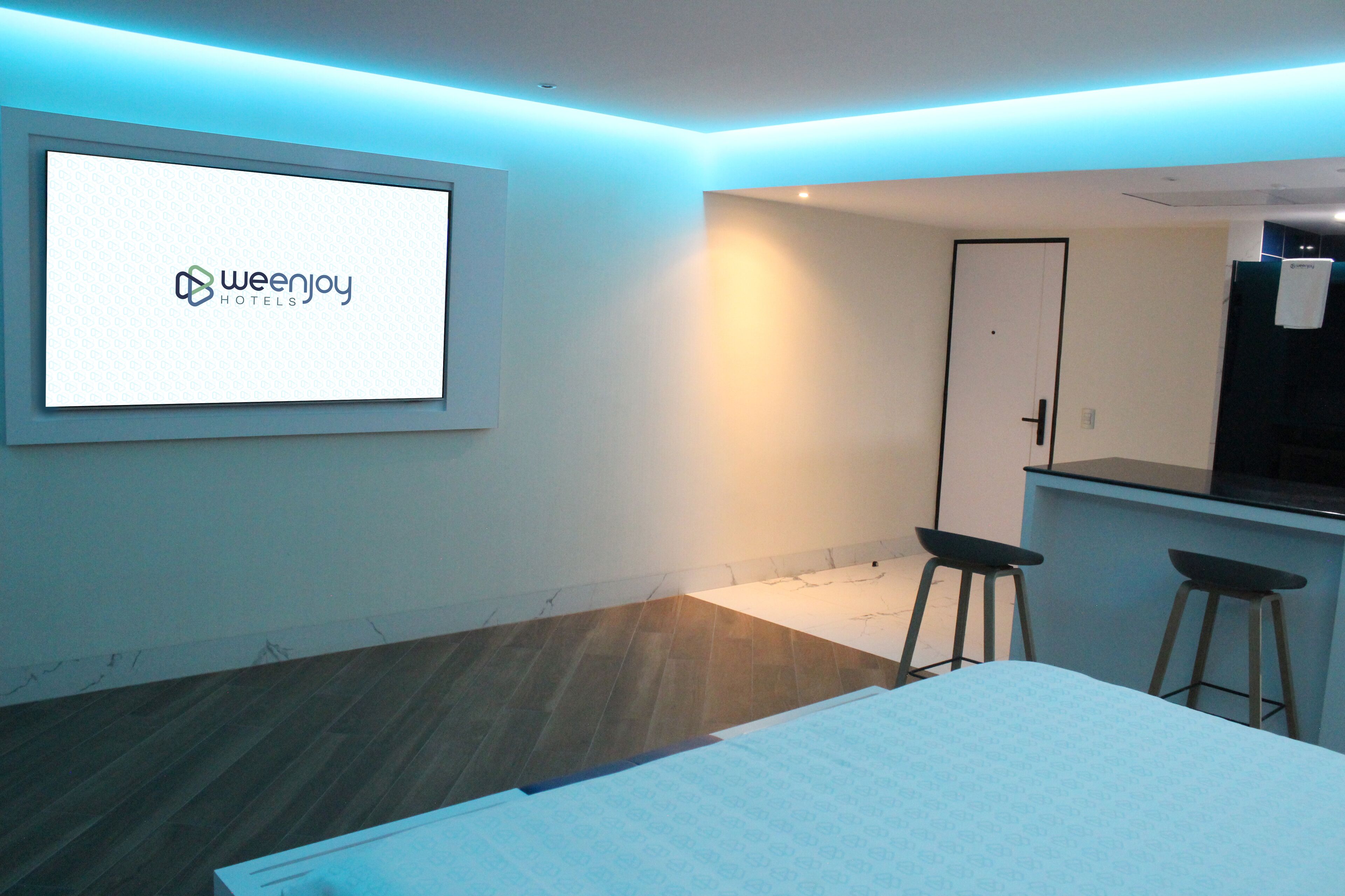 Et moderne hotelværelse med en seng, et skrivebord, to barstole og et stort skærmbillede med Weenjoy Hotels logo.