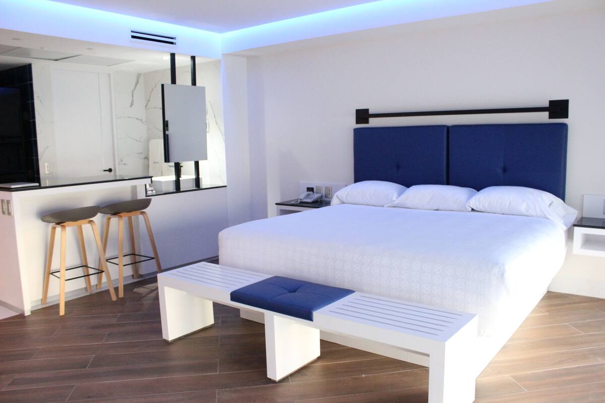 A modern bedroom with a large bed, a bench, two stools, a bedside table, and a mirror.