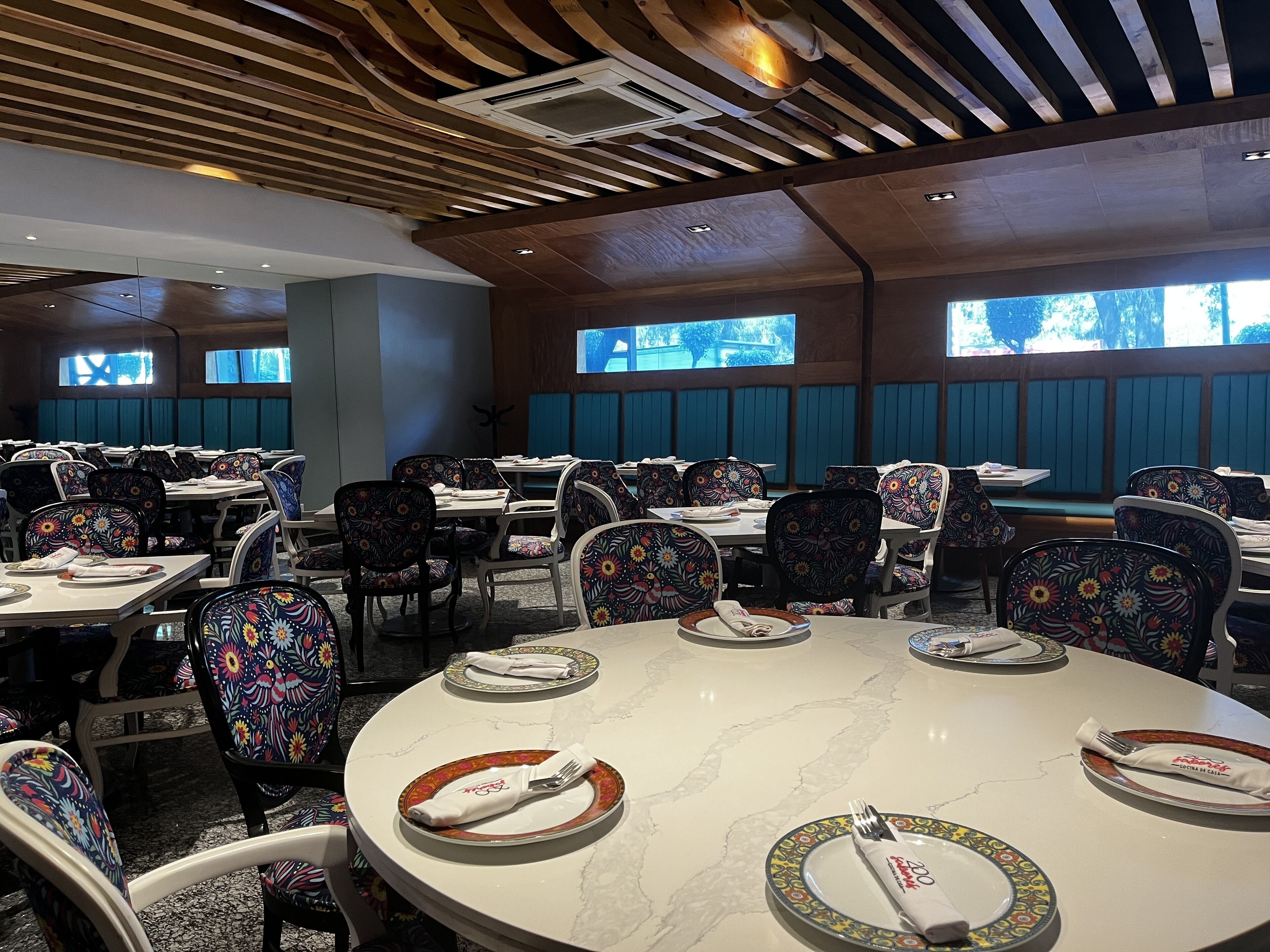 Um restaurante com mesas redondas, cadeiras com padrão floral e teto de madeira.