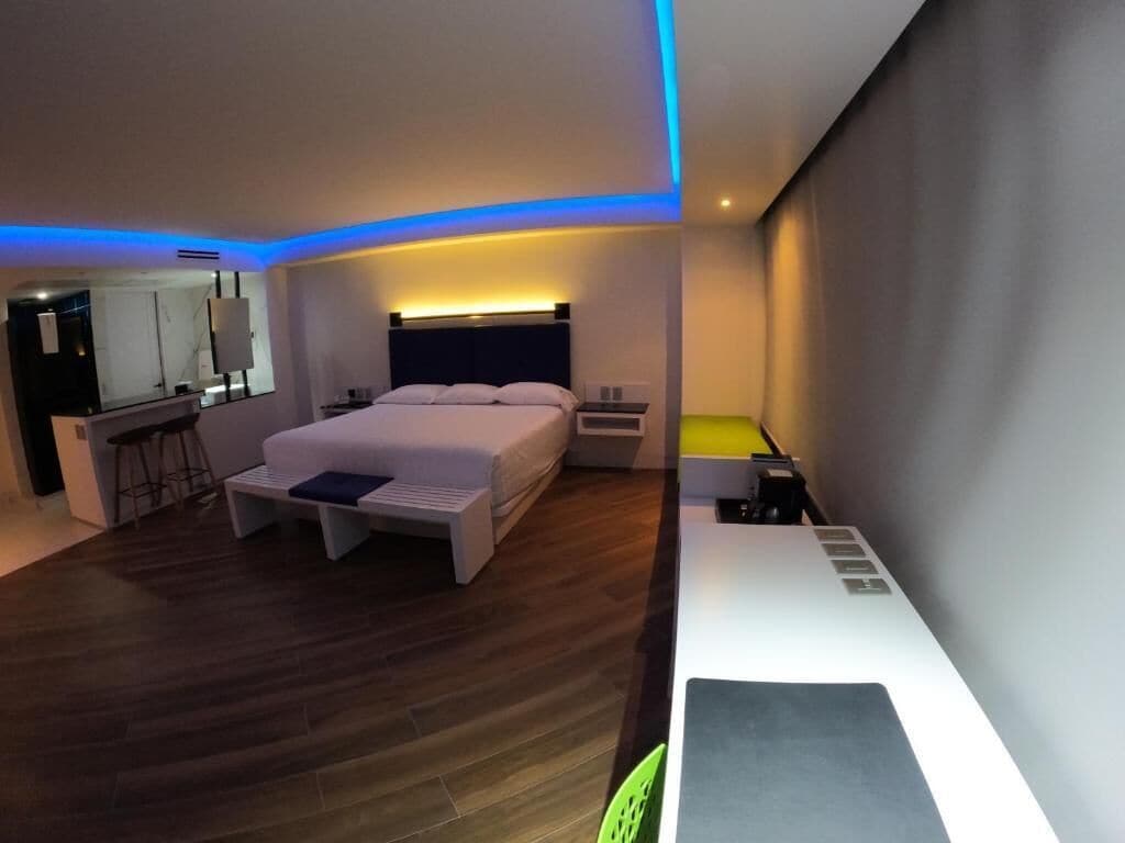 A modern hotel room with a large bed, a desk, and a chair. There is a small table and a bar area in the corner.