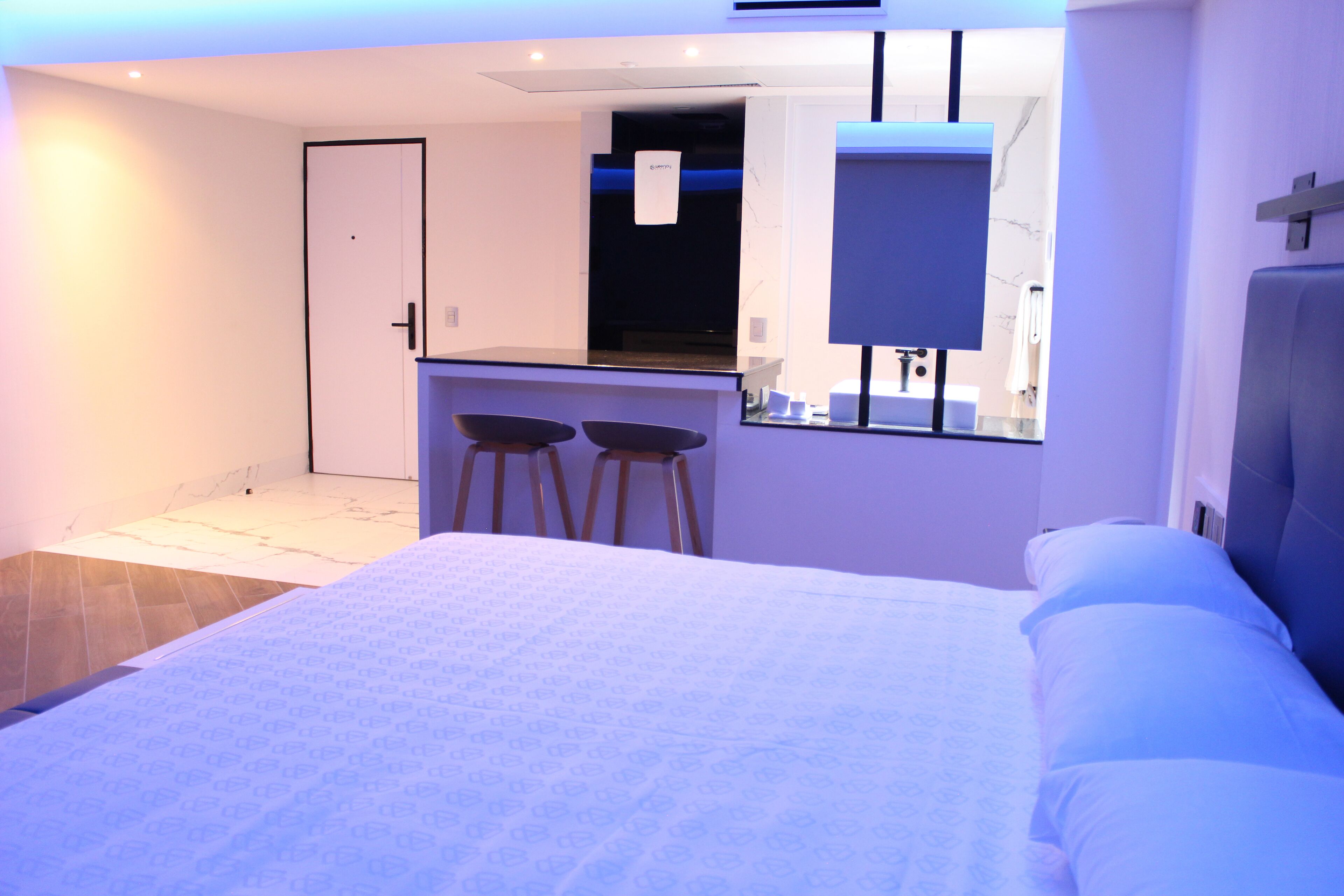 Une chambre d’hôtel moderne dotée d’un grand lit, d’une petite kitchenette et d’une salle de bain visible à travers une paroi en verre.
