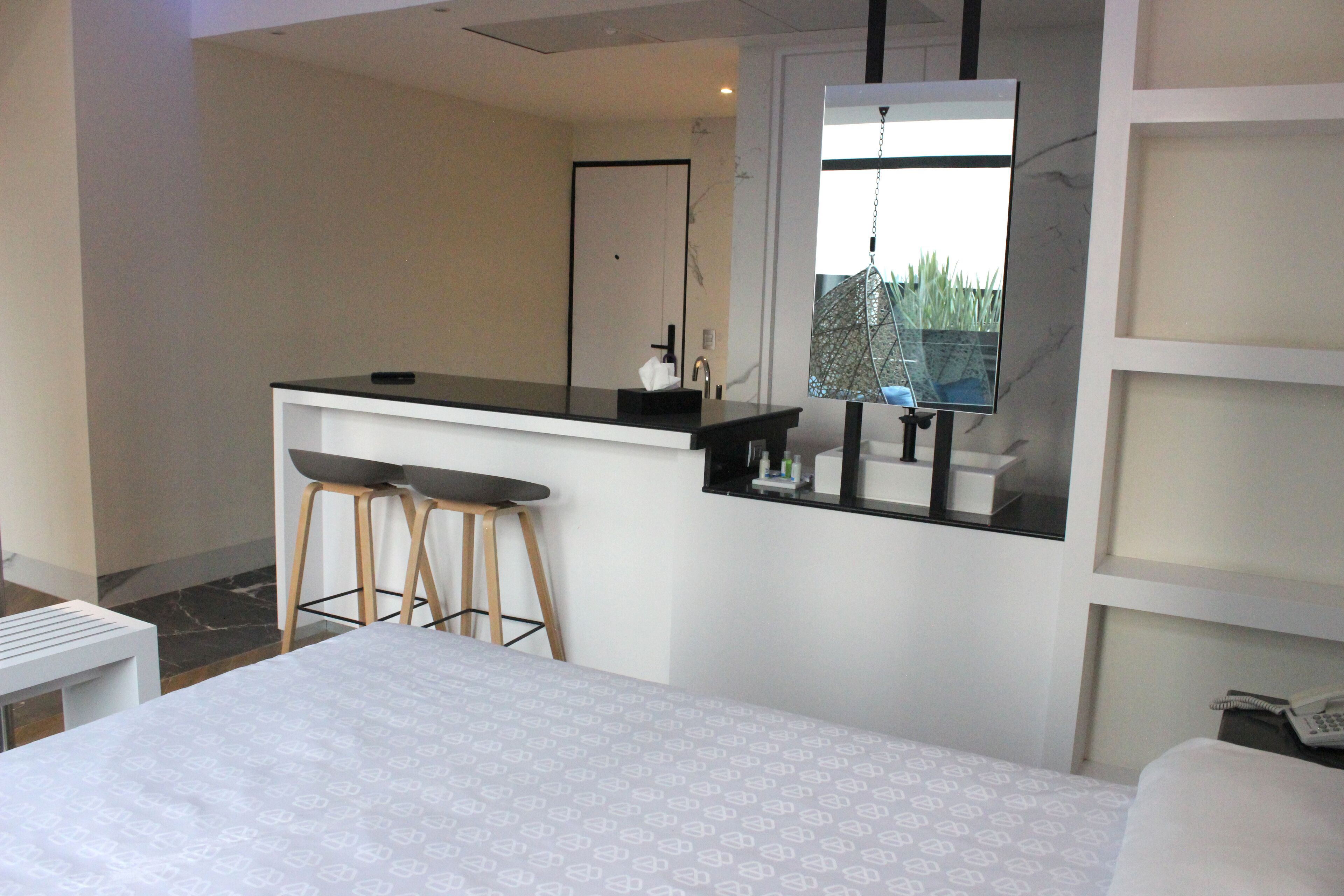 Habitación de hotel con cama, una kitchenette pequeña con taburetes, un espejo y ventana con vista a zonas verdes.
