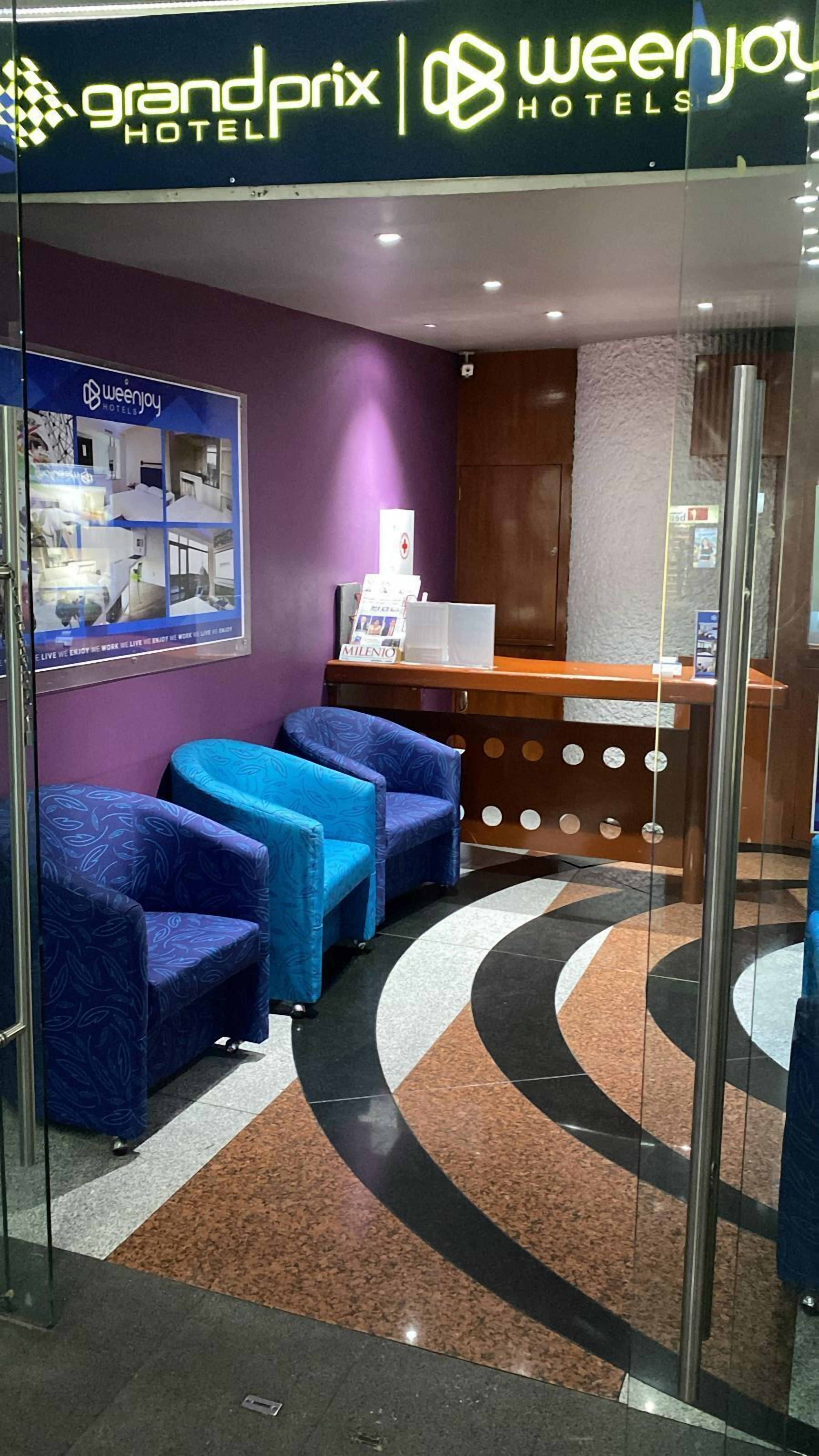A hotel reception area with a purple wall, a reception desk, and three blue armchairs.