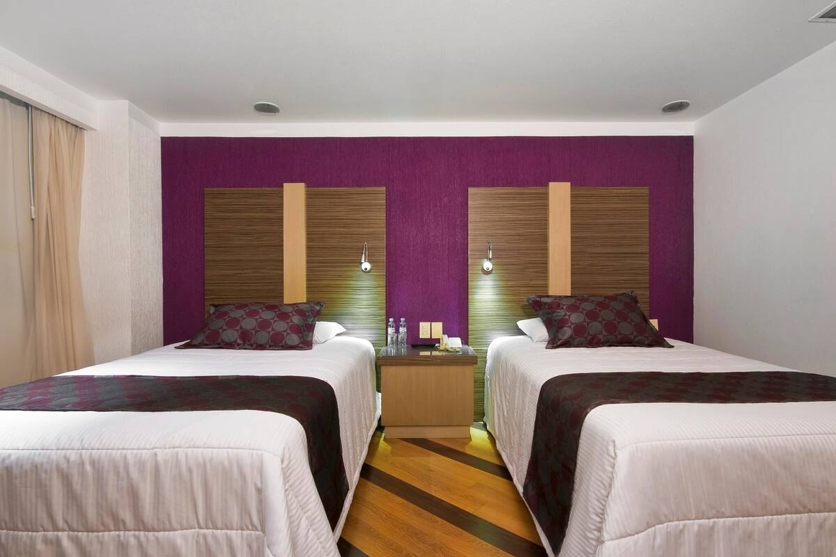 A hotel room with two beds, a wooden floor, a purple accent wall, and a nightstand with a lamp.