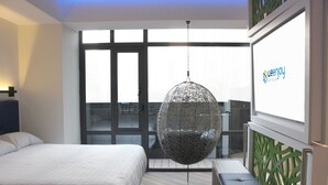 A hotel room with a bed, a hanging chair, a bench, and a large window.