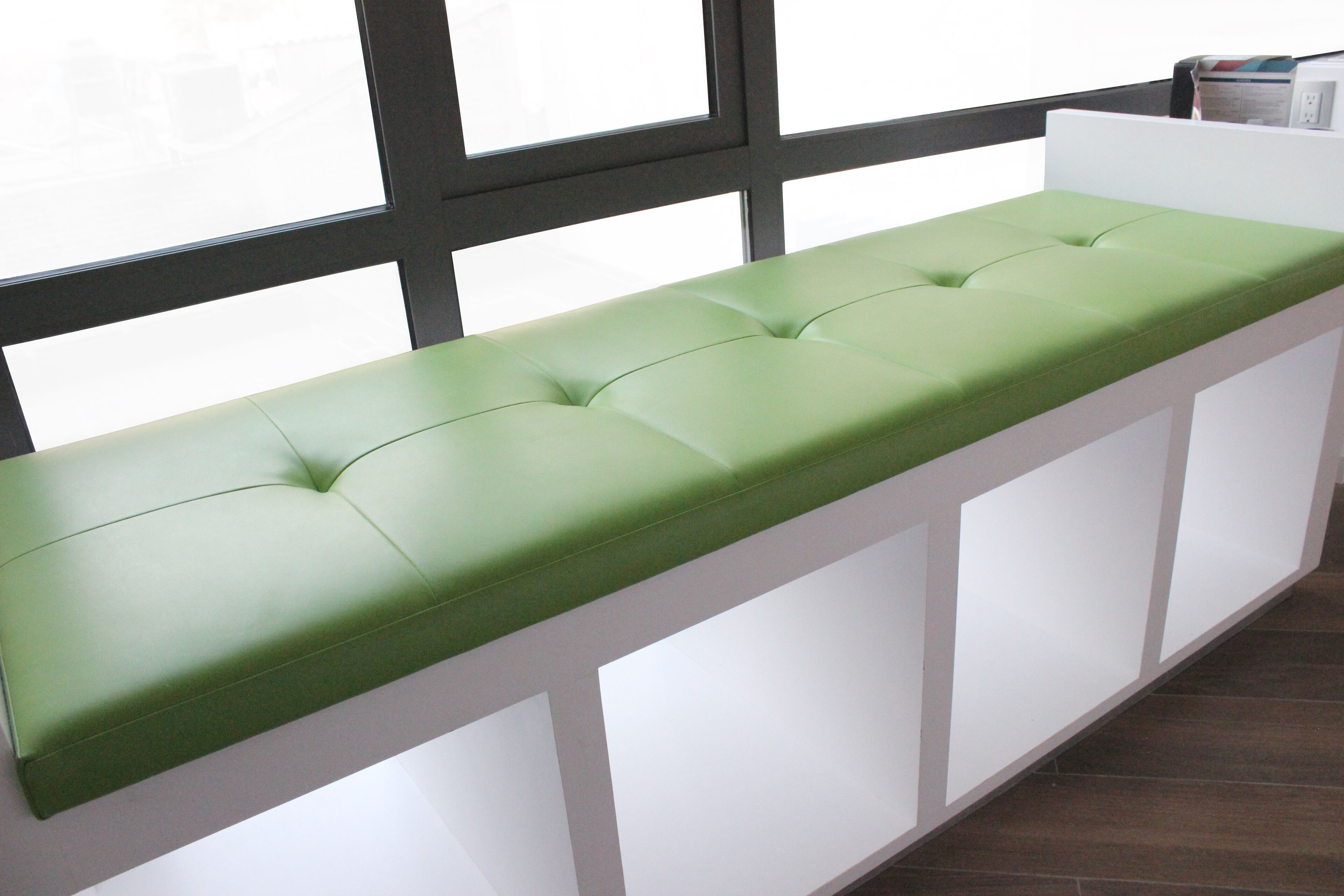 A green cushioned bench with a white base, positioned against a window with a black frame.