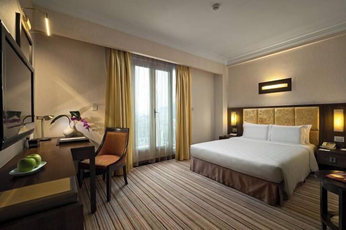 Habitación de hotel con cama, escritorio con silla, espejo, mesita de noche y ventana con cortinas.