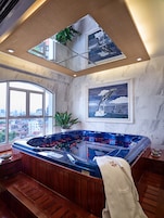 A modern bathroom with a large bathtub, wooden bench, potted plants, and city views through large windows.