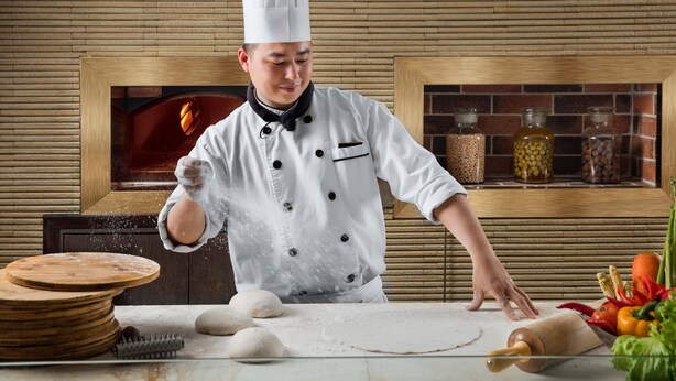 Un chef, vêtu d’un uniforme blanc et coiffé d’un haut-de-forme, prépare une pâte sur un plan de travail de cuisine, en utilisant divers ingrédients et ustensiles.