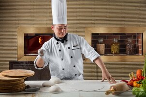 A chef in a white uniform with a tall hat, preparing dough on a kitchen counter with various ingredients and tools.