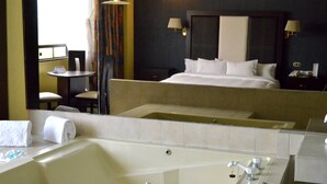 Quarto de hotel com uma banheira grande, uma cama com roupa de cama branca, uma pequena mesa redonda com uma cadeira e uma mesinha de cabeceira com abajur.
