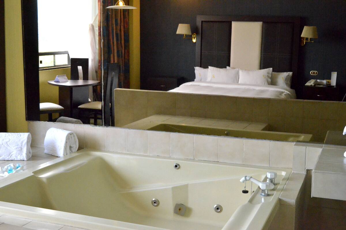 A hotel room with a large bathtub, a bed with white linens, a small round table with a chair, and a nightstand with a lamp.
