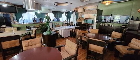Un restaurante con mesas redondas y sillas acolchadas, una zona de cocina y una pared verde con ventanas.