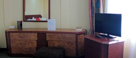 A hotel room with a wooden vanity, a mirror, a television on a stand, and a small ottoman.
