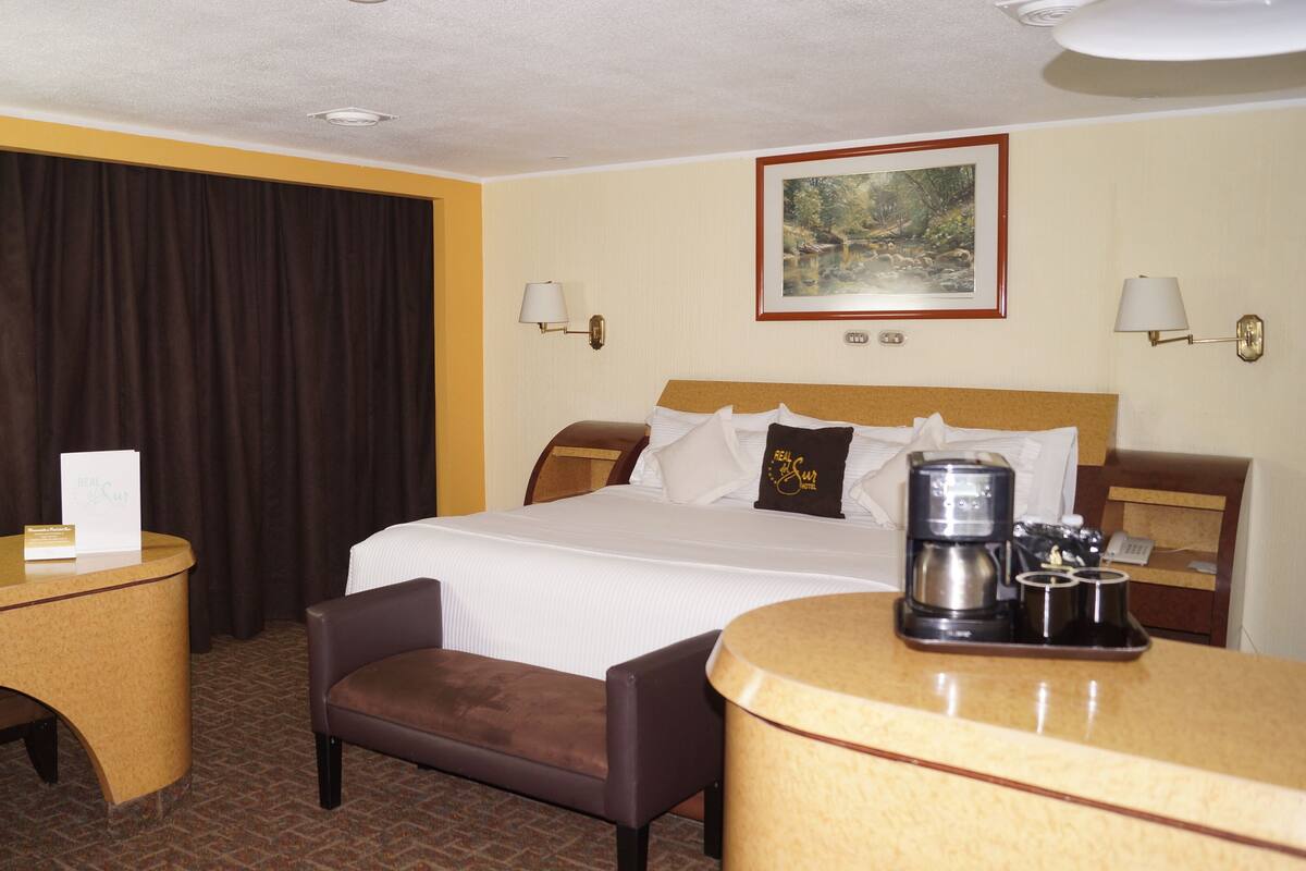 A hotel room with a bed, a desk, a chair, a coffee maker, and a painting on the wall.