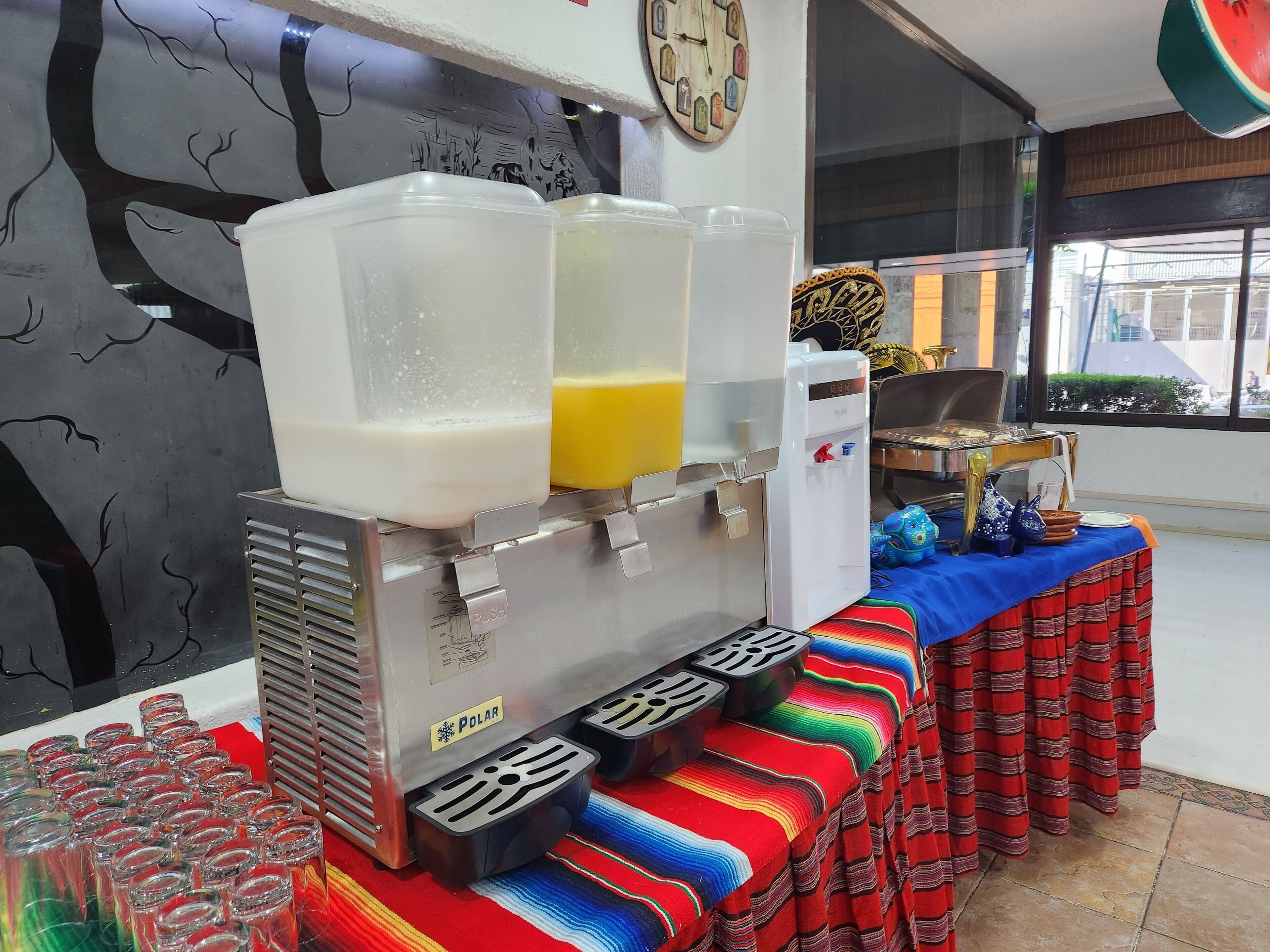 Un área de bebidas con dispensador de agua, dispensador de jugo, vasos y una mesa con mantel colorido.