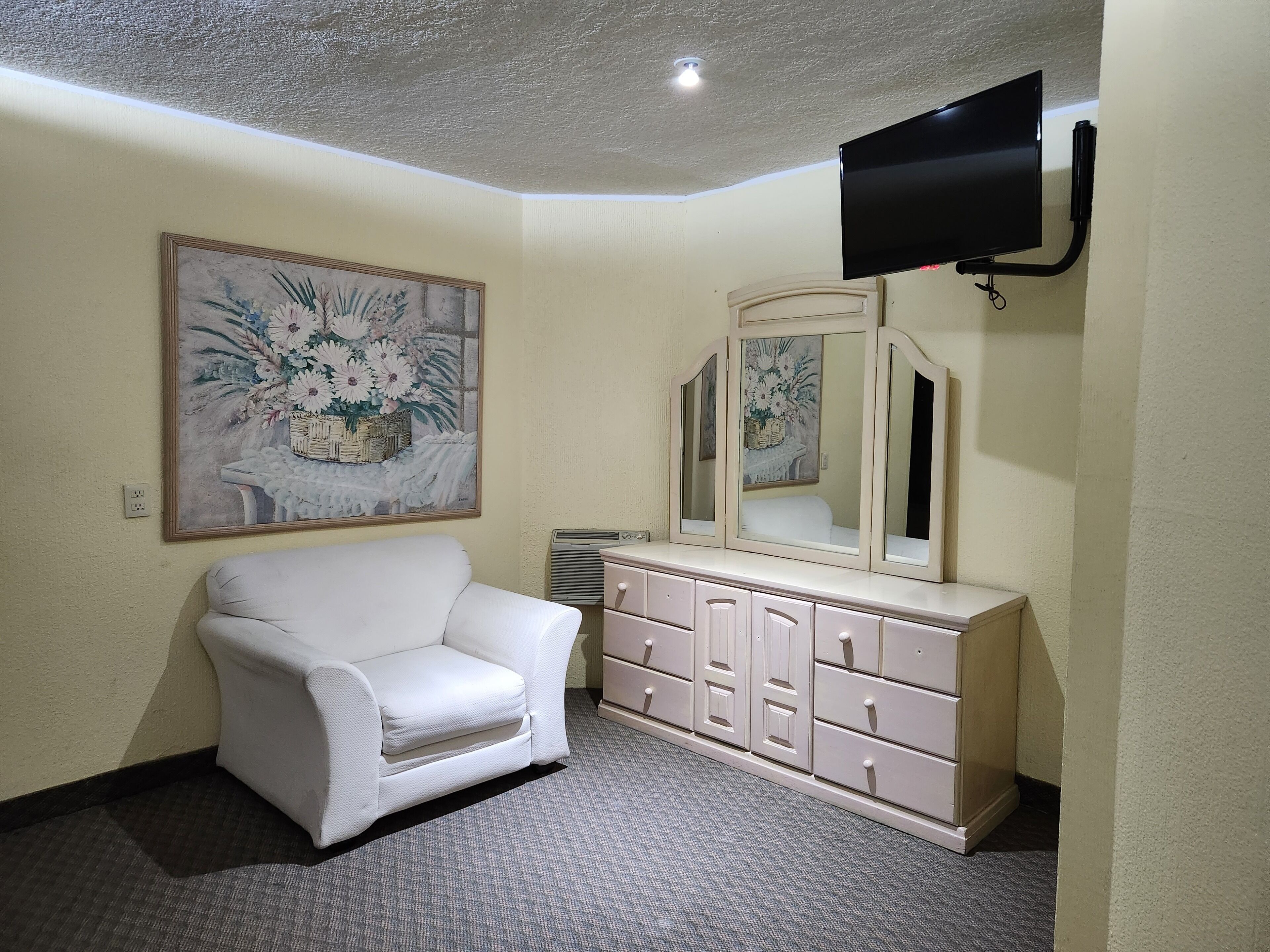 A room with a white armchair, a dresser with drawers, a large mirror, and a flat-screen TV mounted on the wall.