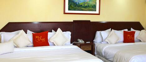 A hotel room with two beds, each with white bedding and red pillows. There is a painting of a mountain landscape on the wall above the beds.