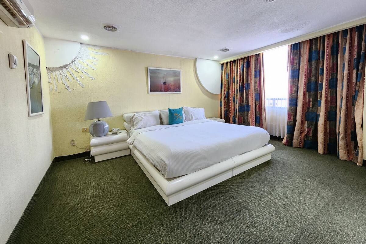 A hotel room with a large bed, a bedside table with a lamp, a phone, and a wall-mounted air conditioner.