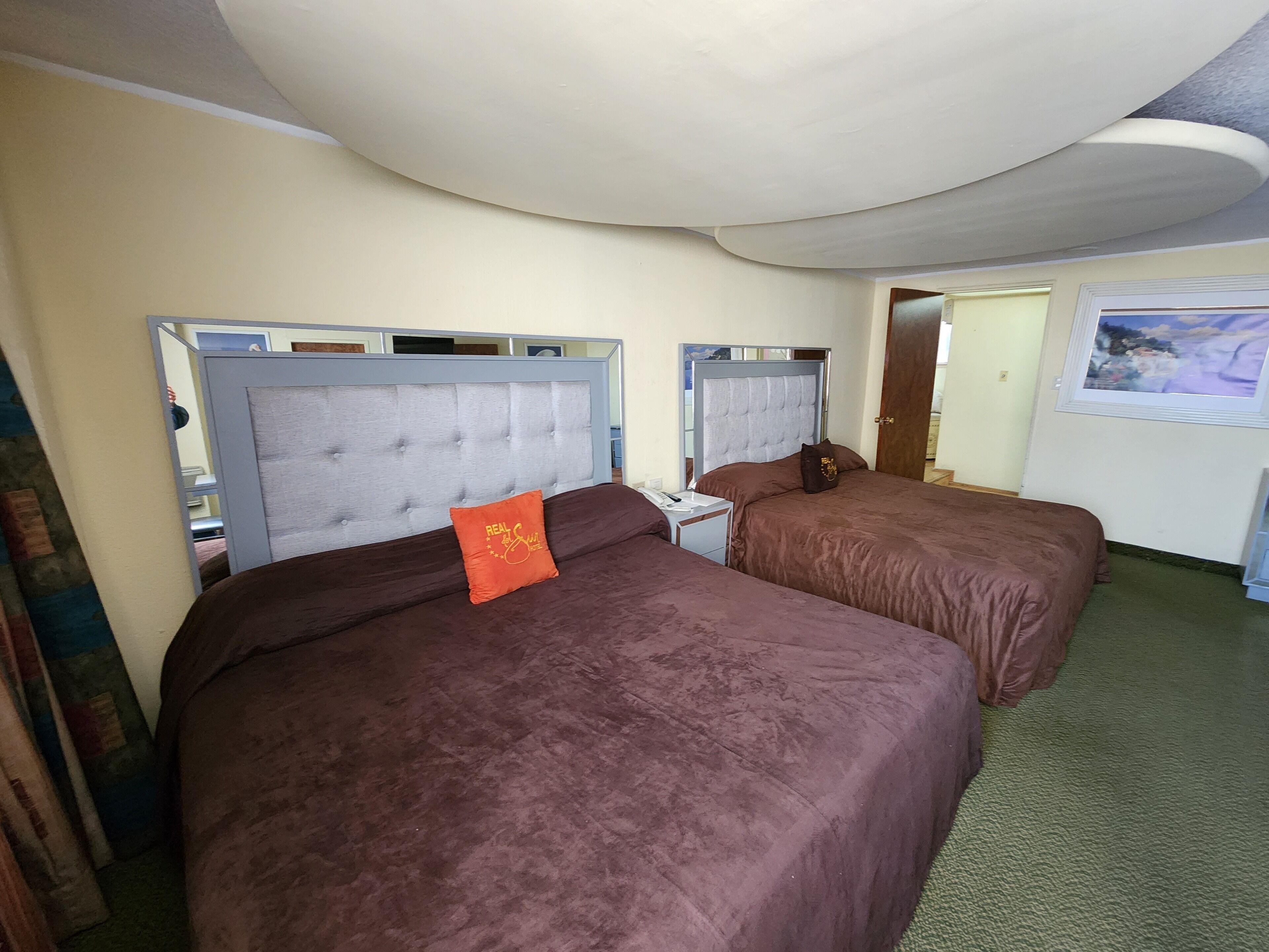 Ein Hotelzimmer mit zwei Betten, jeweils mit Kopfteil und Kissen. Ein Nachttisch mit Lampe und kleinem Tisch sind vorhanden. Der Raum hat einen grünen Teppich und ein Bild an der Wand.