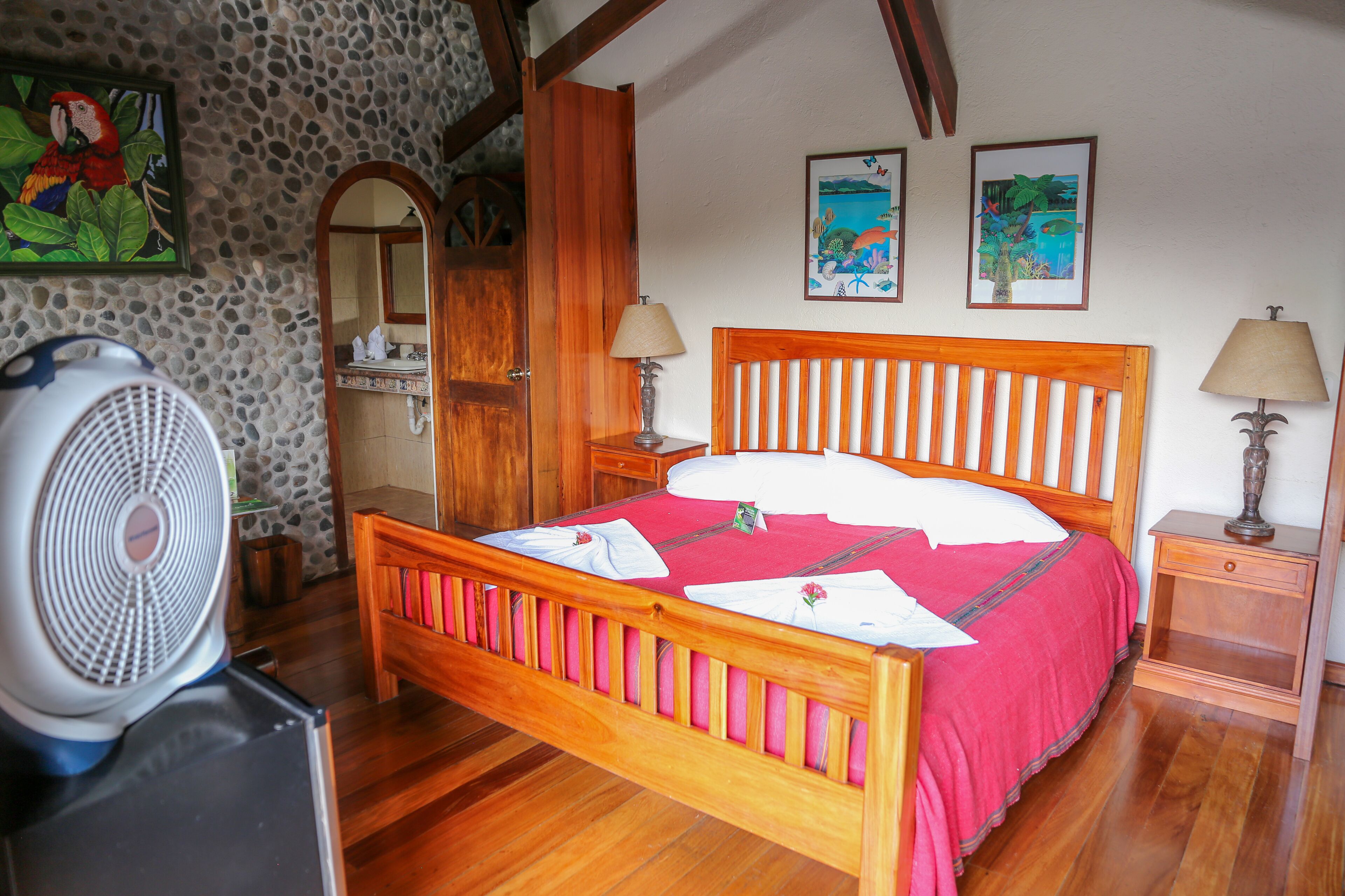 A wooden bed with a red bedspread, two bedside tables with lamps, a mirror, and a fan.
