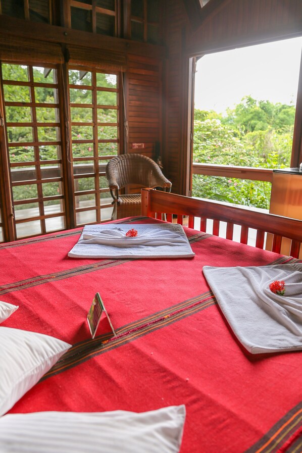 Una habitación con una cama cubierta con una manta roja, una silla de madera y vistas a la vegetación a través de amplias ventanas.