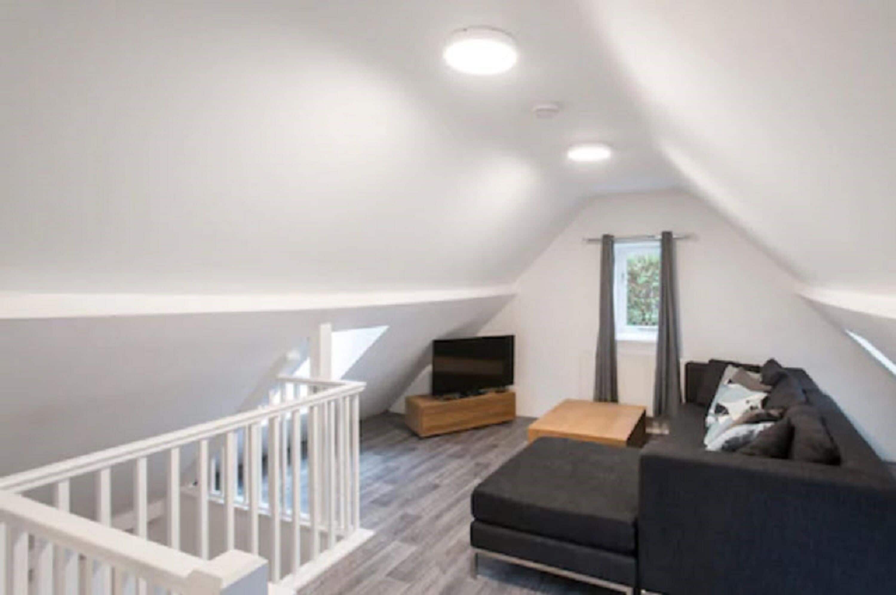 A modern loft apartment with a high ceiling, a wooden entertainment unit, a black sofa, and a staircase with white railings.